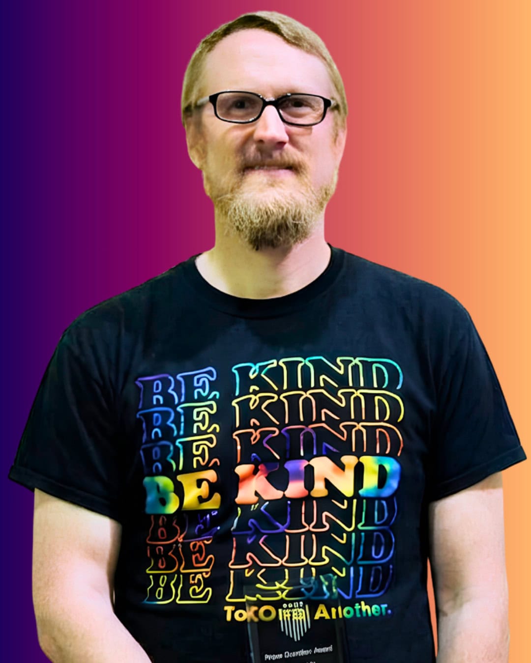 DennyHam A man with glasses and a beard wears a black “BE KIND” t-shirt with a rainbow effect, standing against a gradient orange and purple background.