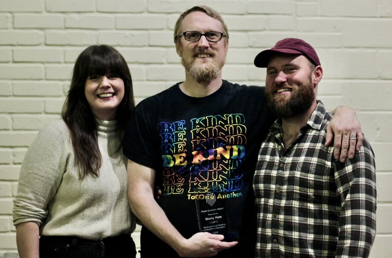 DennyHam Three people stand in front of a white brick wall; the person in the center holds a glass award and wears a “Be Kind” shirt, with the other two smiling beside them.