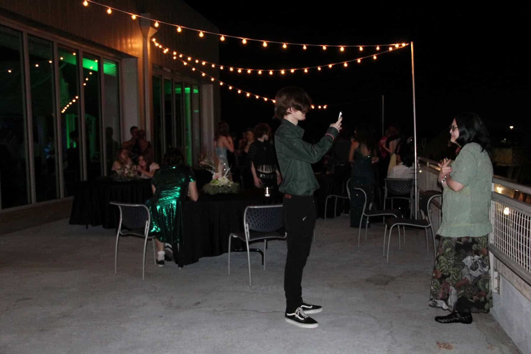 A person takes a photo of another person at a night-time outdoor event with string lights and attendees in the background.