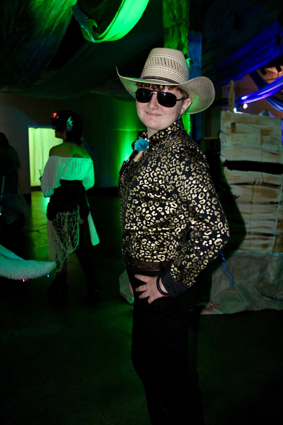 A person wearing a leopard print shirt, sunglasses, and a straw hat stands posing with a hand on their hip at a party with green lighting in the background.