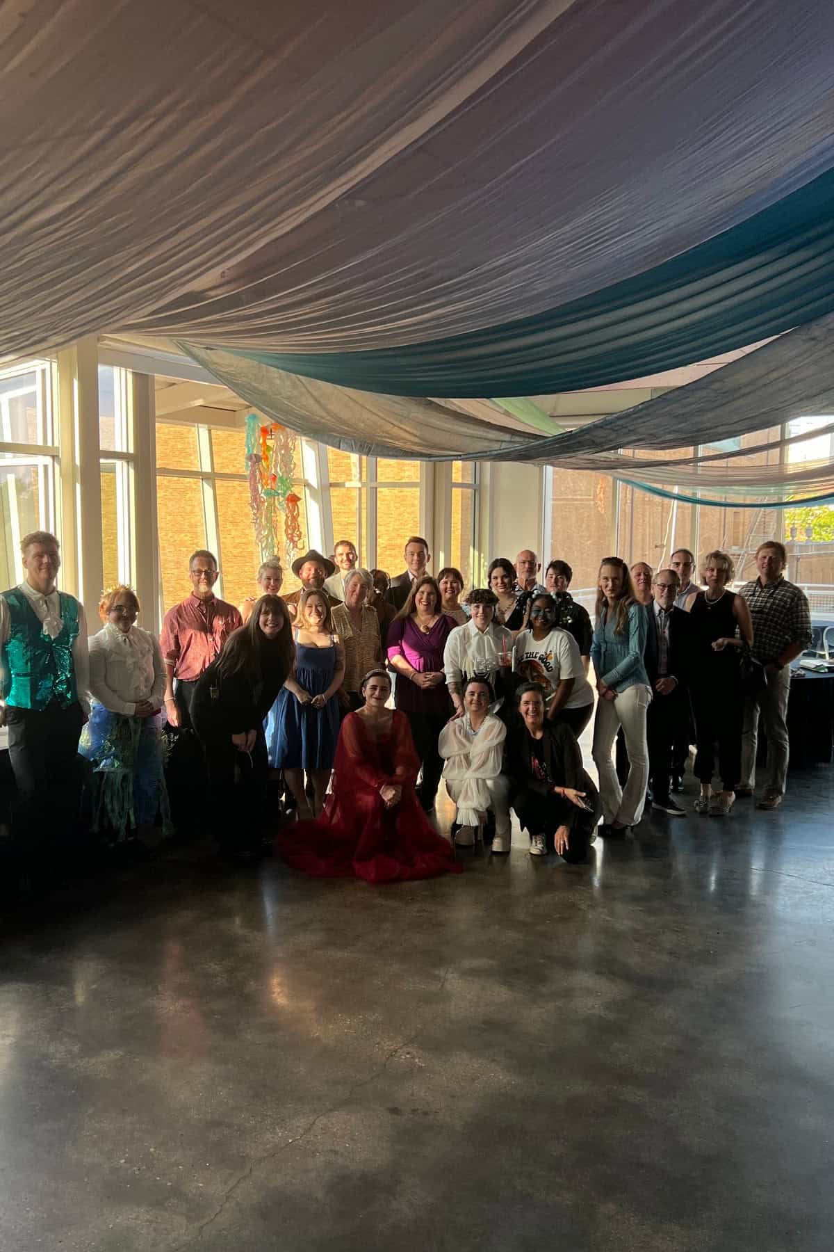 A group of people dressed in various colorful outfits posing for a photo in a room with draped ceilings and large windows.