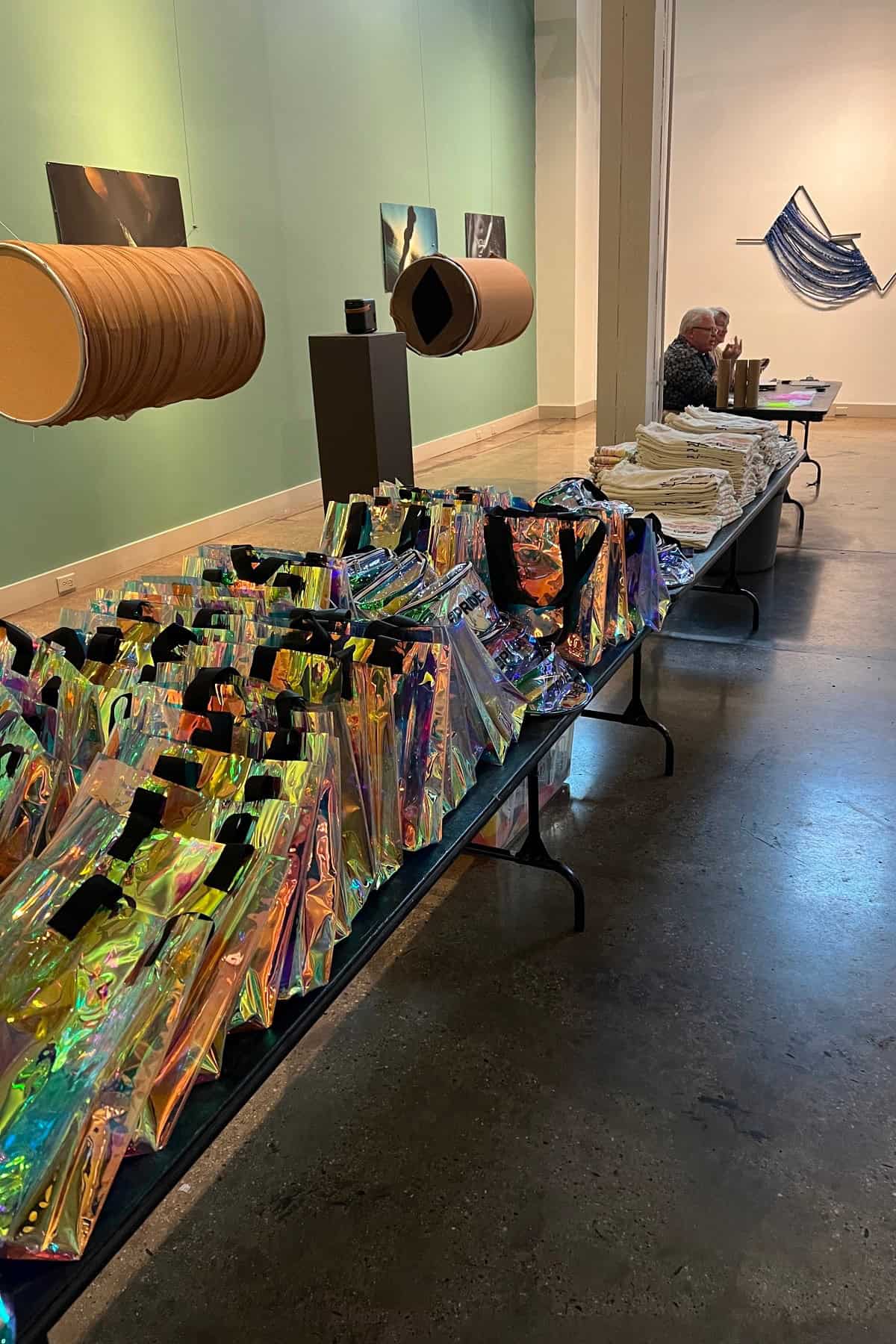 An art gallery with iridescent bags on a table in the foreground, cylindrical objects on stands, and an elderly person sitting at a table with stacked papers in the background.