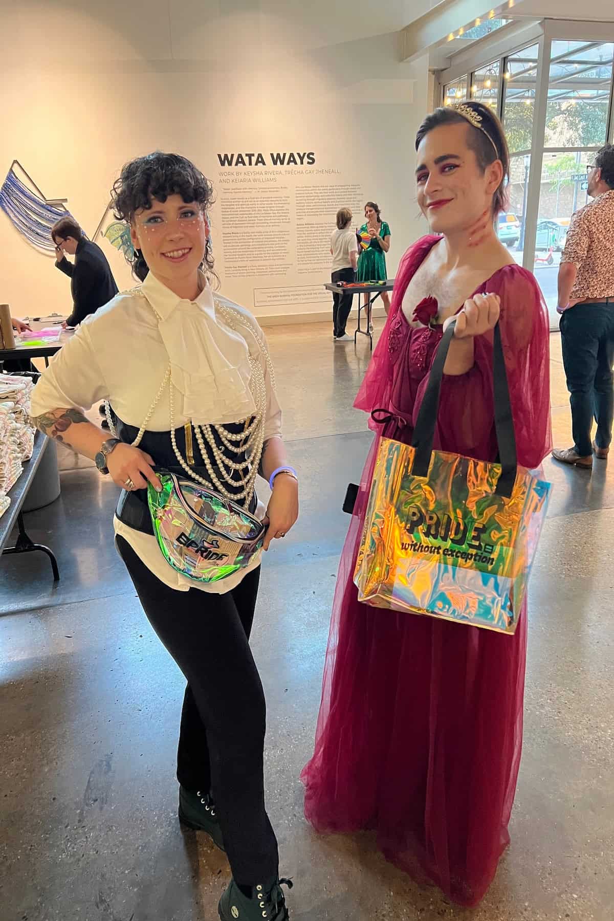 Two people pose indoors holding holographic bags. One wears a white blouse and black pants, the other a red dress. Art installation visible in the background.