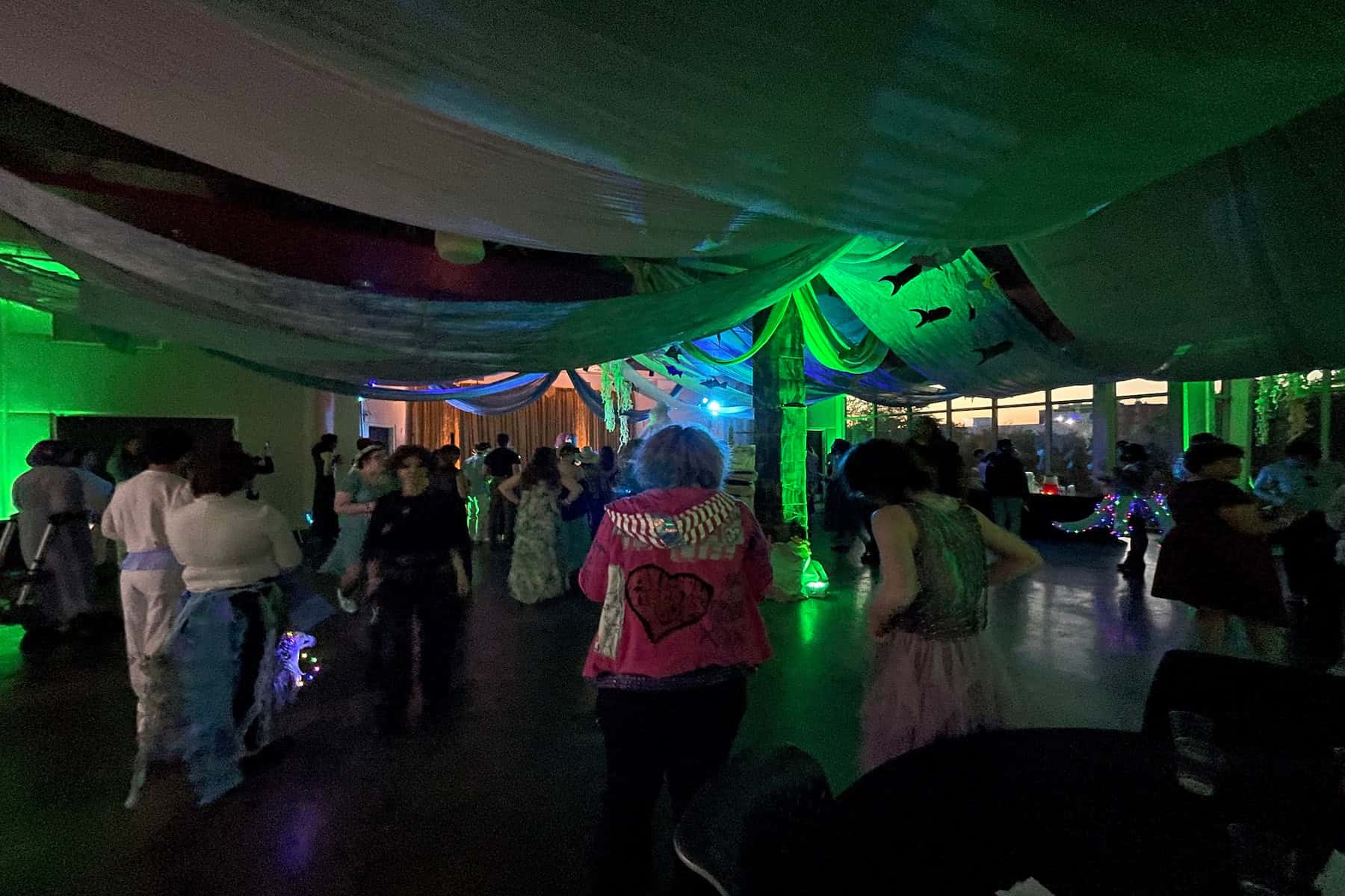 People dancing at a dimly lit party with colorful lights and draped ceiling decorations.