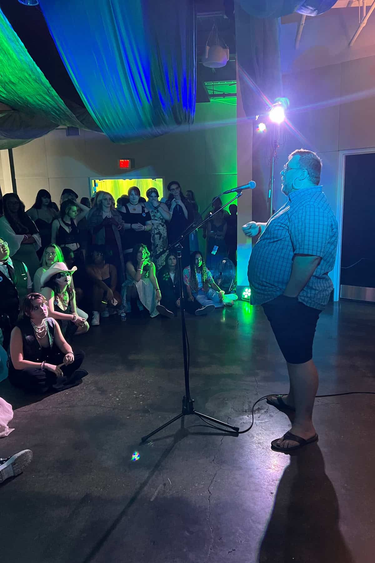 A person speaking into a microphone on a stage at a dimly lit event with an audience watching in the background.