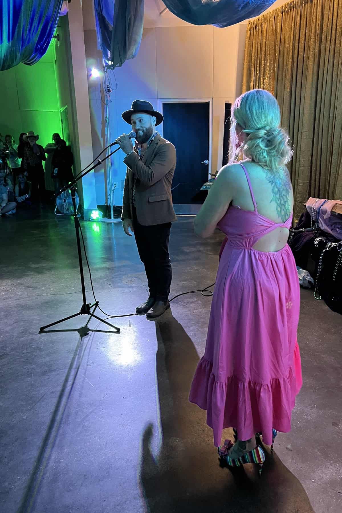 A man in a hat speaking into a microphone at an event, facing a woman in a pink dress who is listening attentively.