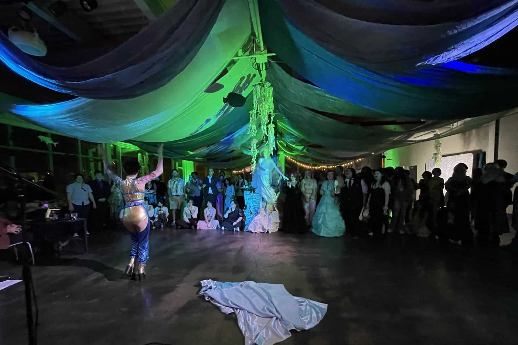 A drumming performer playing in front of an audience under a canopy of draped blue and green fabrics in a dimly lit event space.