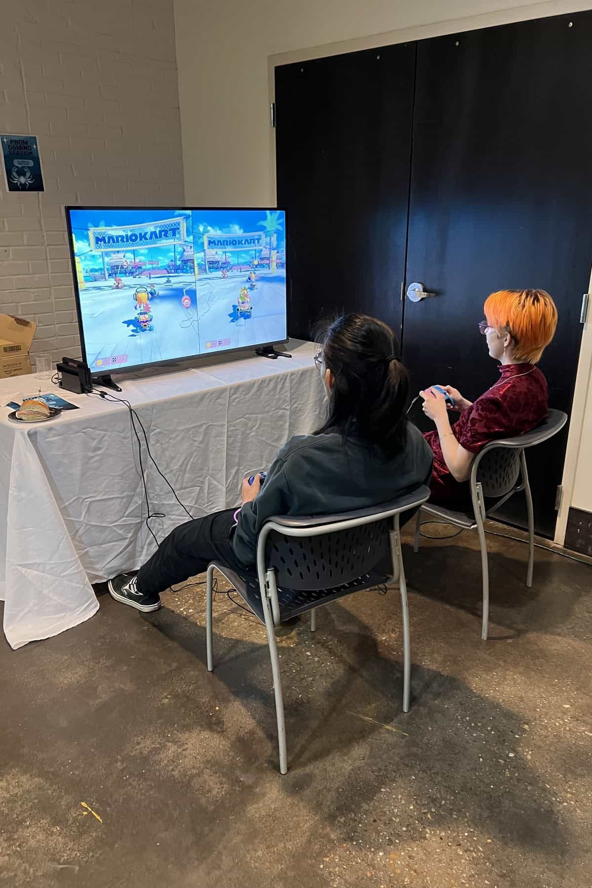 Two people playing Mario Kart on a console connected to a large screen in a casual indoor setting.