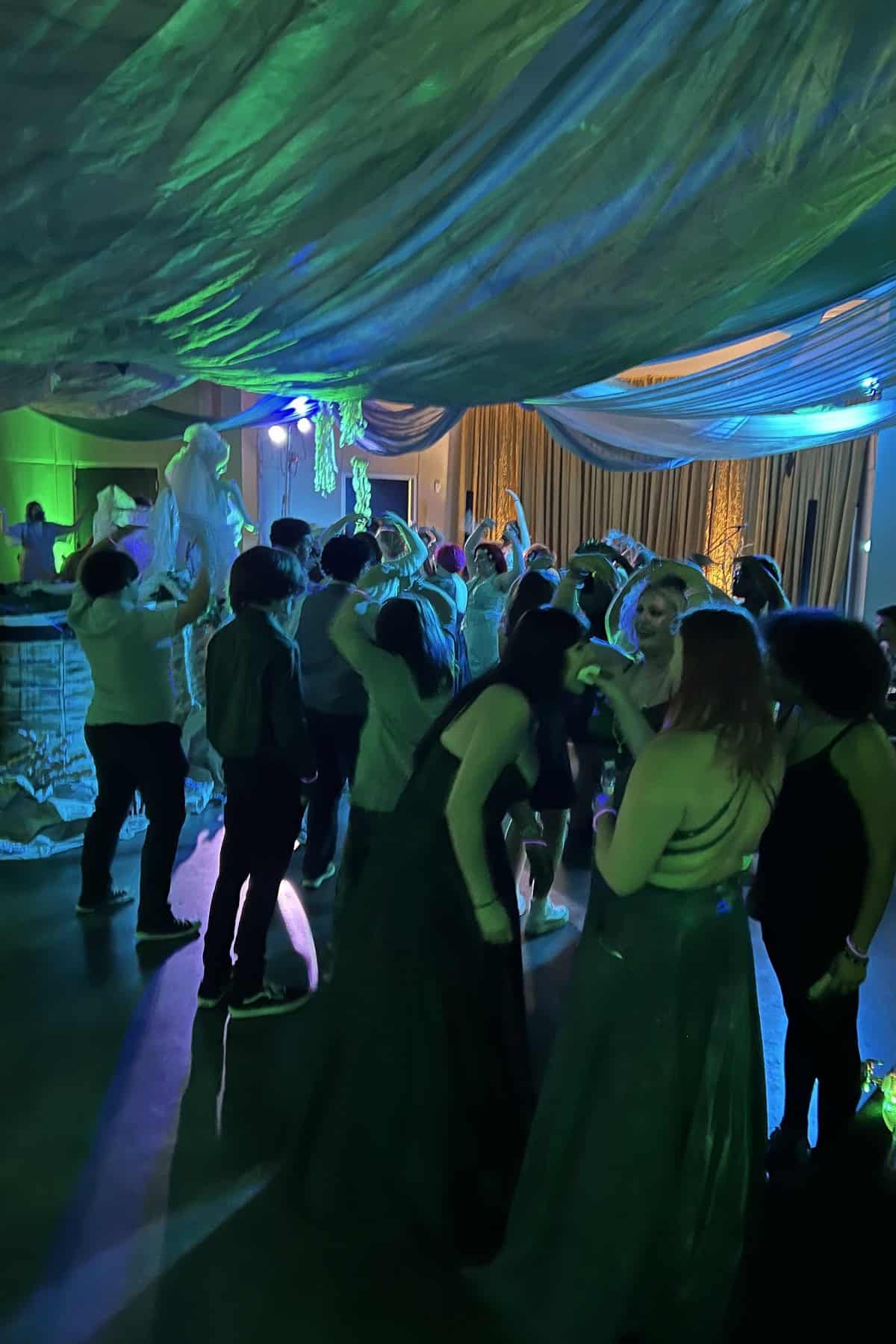 Group of people dancing under green lights at an indoor party with draped ceiling decorations.