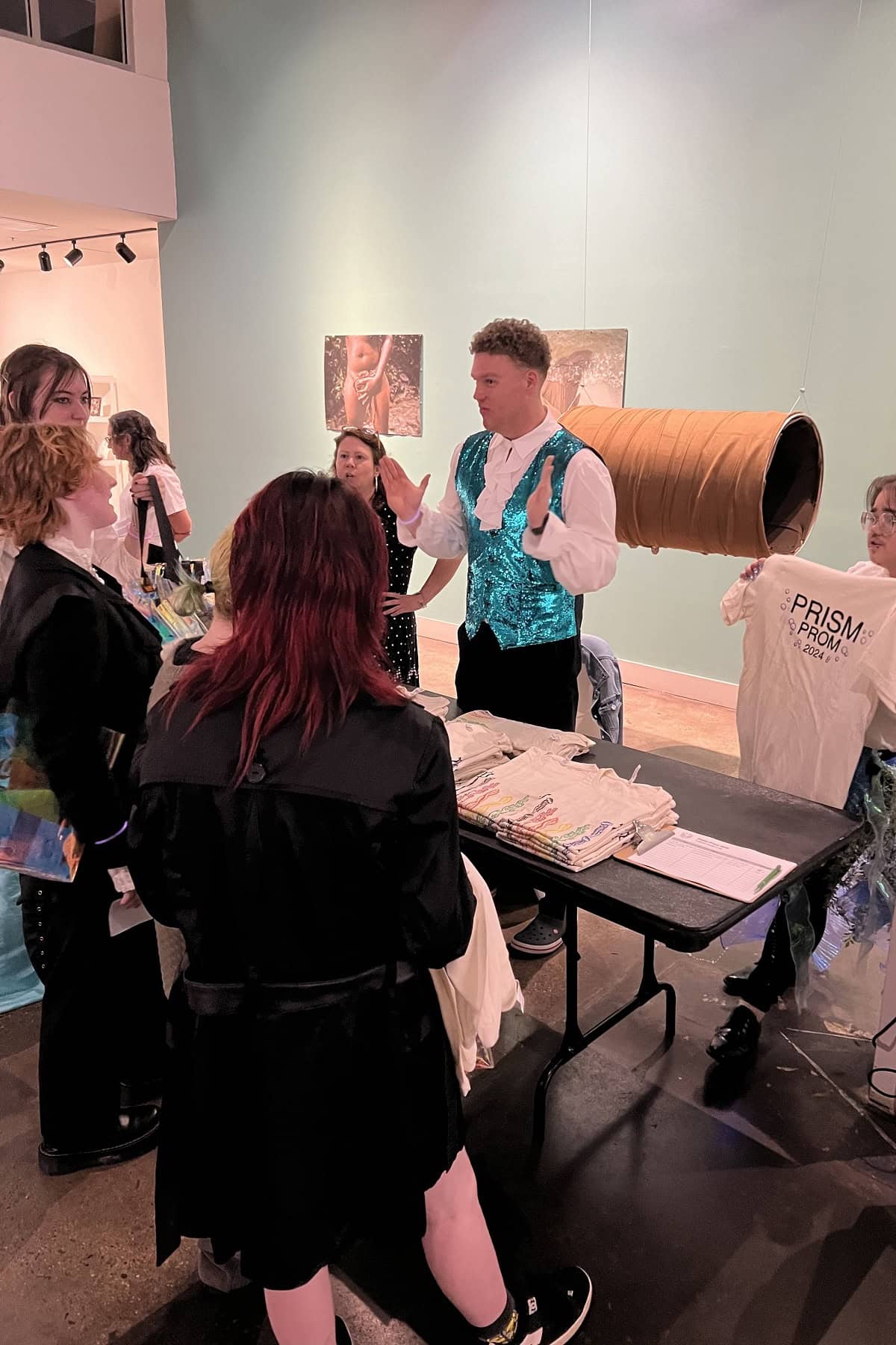 A man in a blue sequined vest demonstrates a large cylindrical object to a group of people at an indoor exhibition.