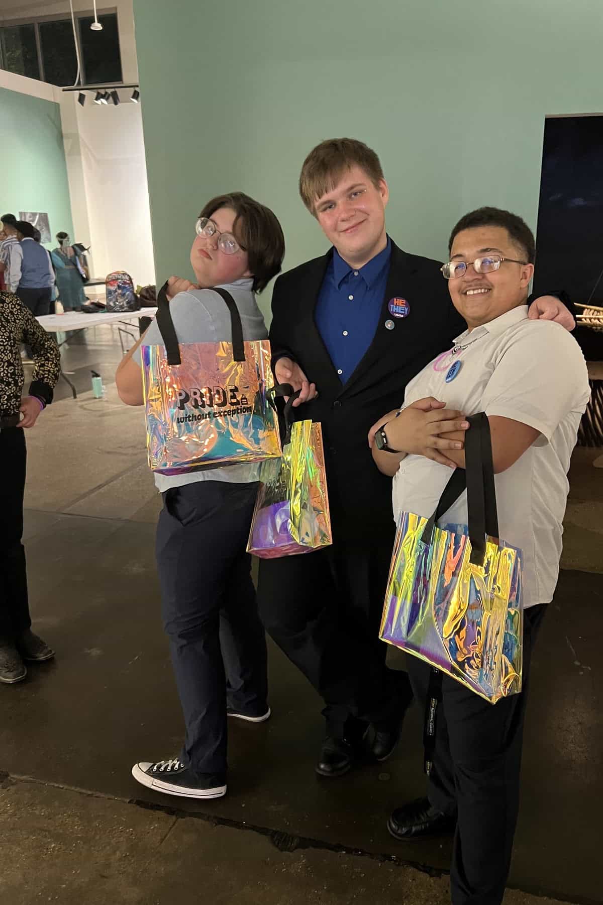 Three people smiling at an event, holding colorful tote bags with "Pride" written on them. They are standing closely together, posing for the photo.