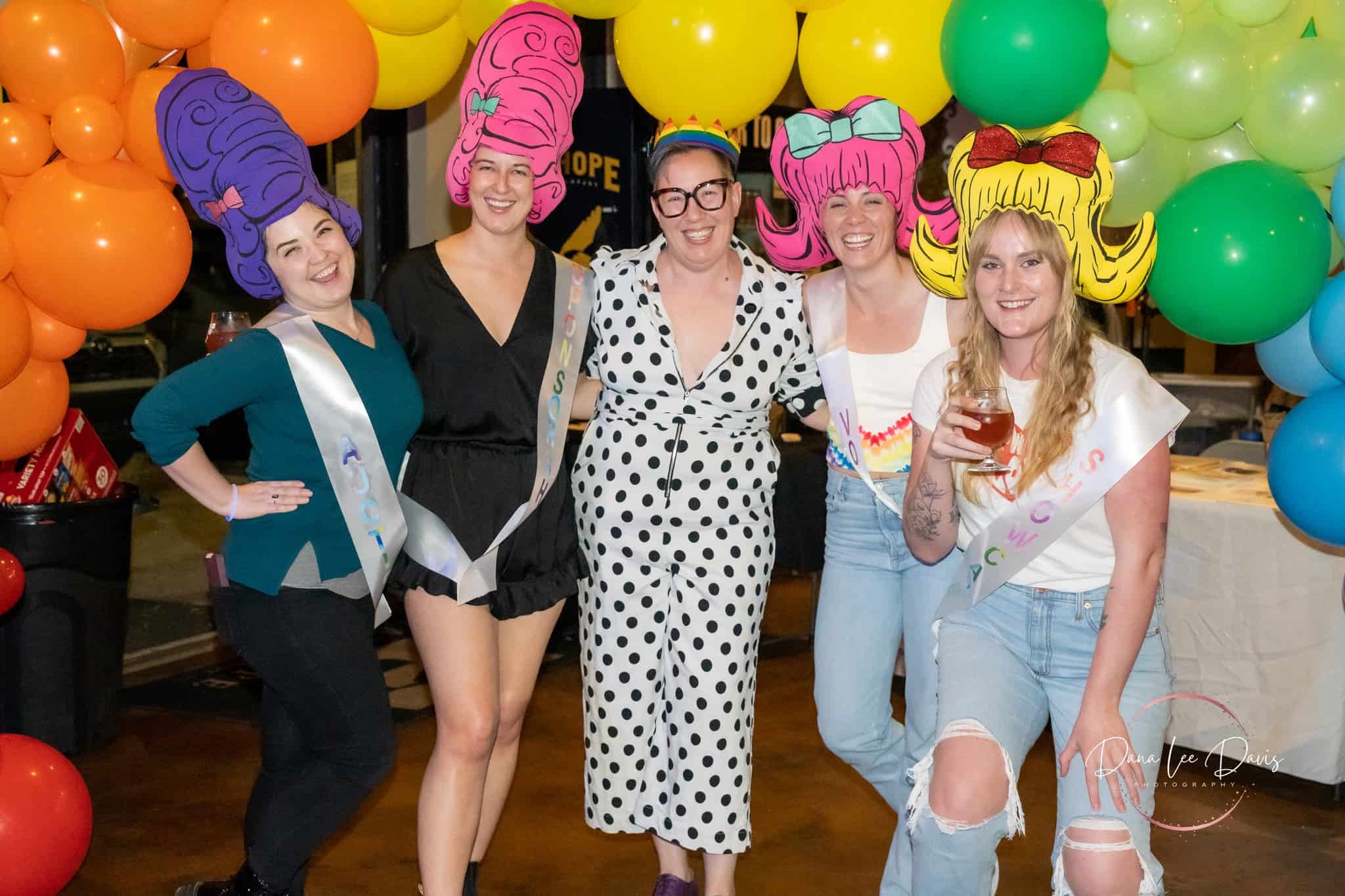 Group of smiling people at a party wearing colorful whimsical wigs and party attire, with balloons in the background.