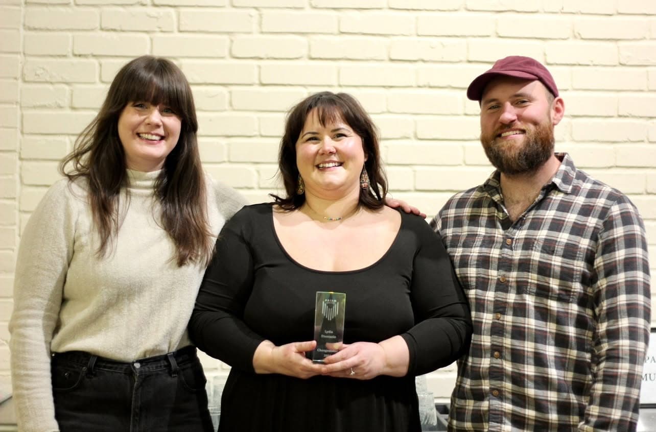 LydiaShantazio Three people stand in front of a white brick wall, smiling. The person in the center holds a small trophy or award.