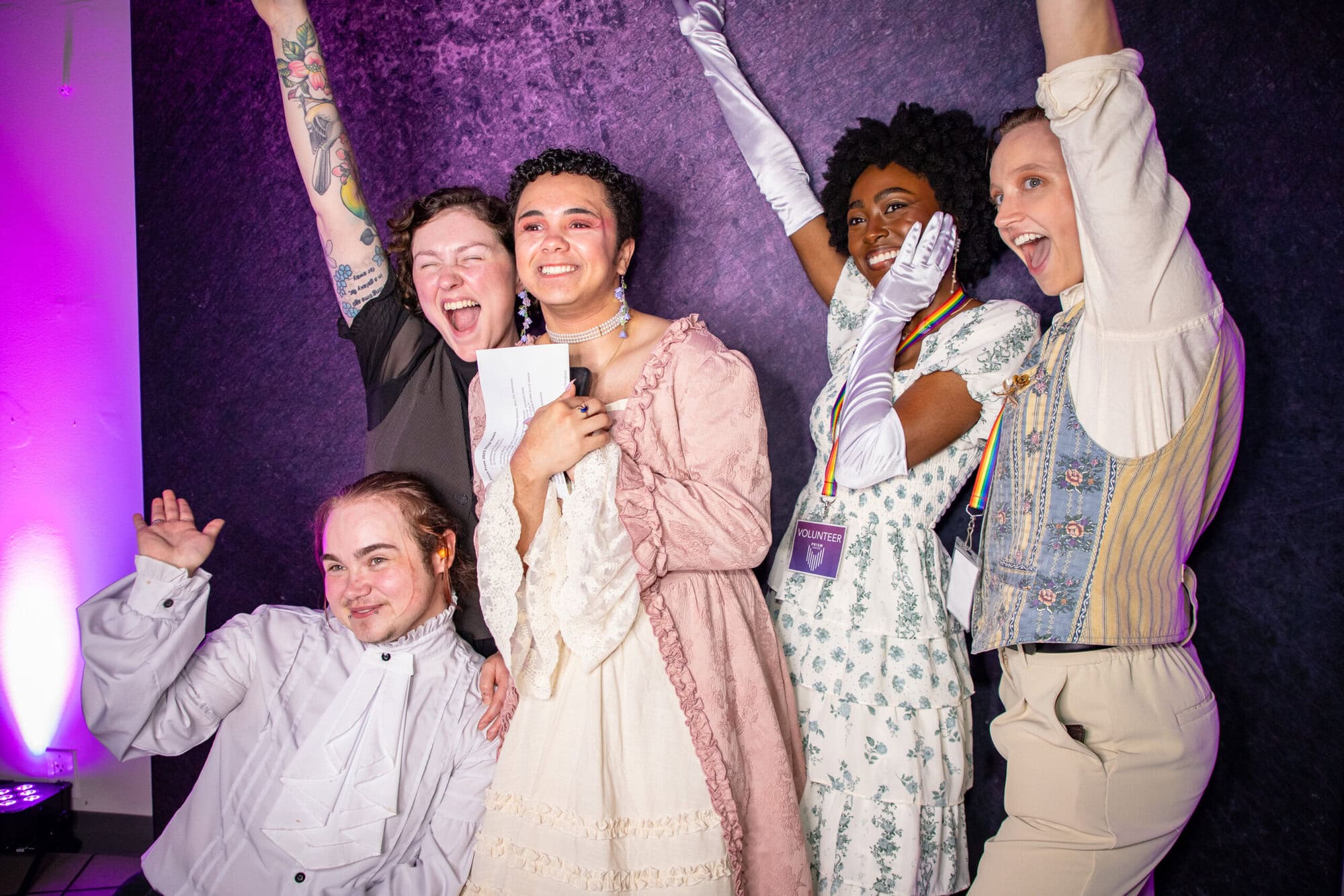 Five people in various costumes pose together, smiling and raising their arms, in front of a purple textured backdrop with pink lighting on the side.