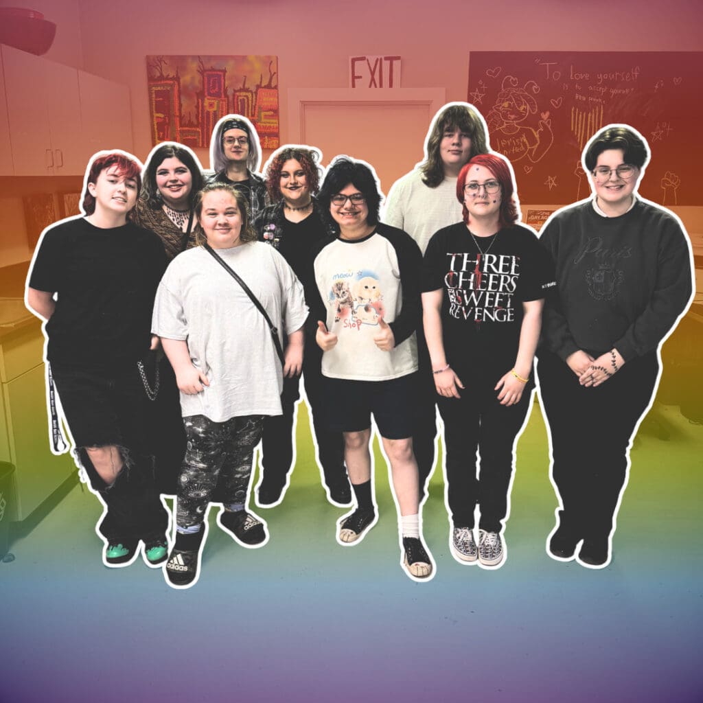 A group of nine people stand together in a brightly lit room with colorful walls and drawings in the background.