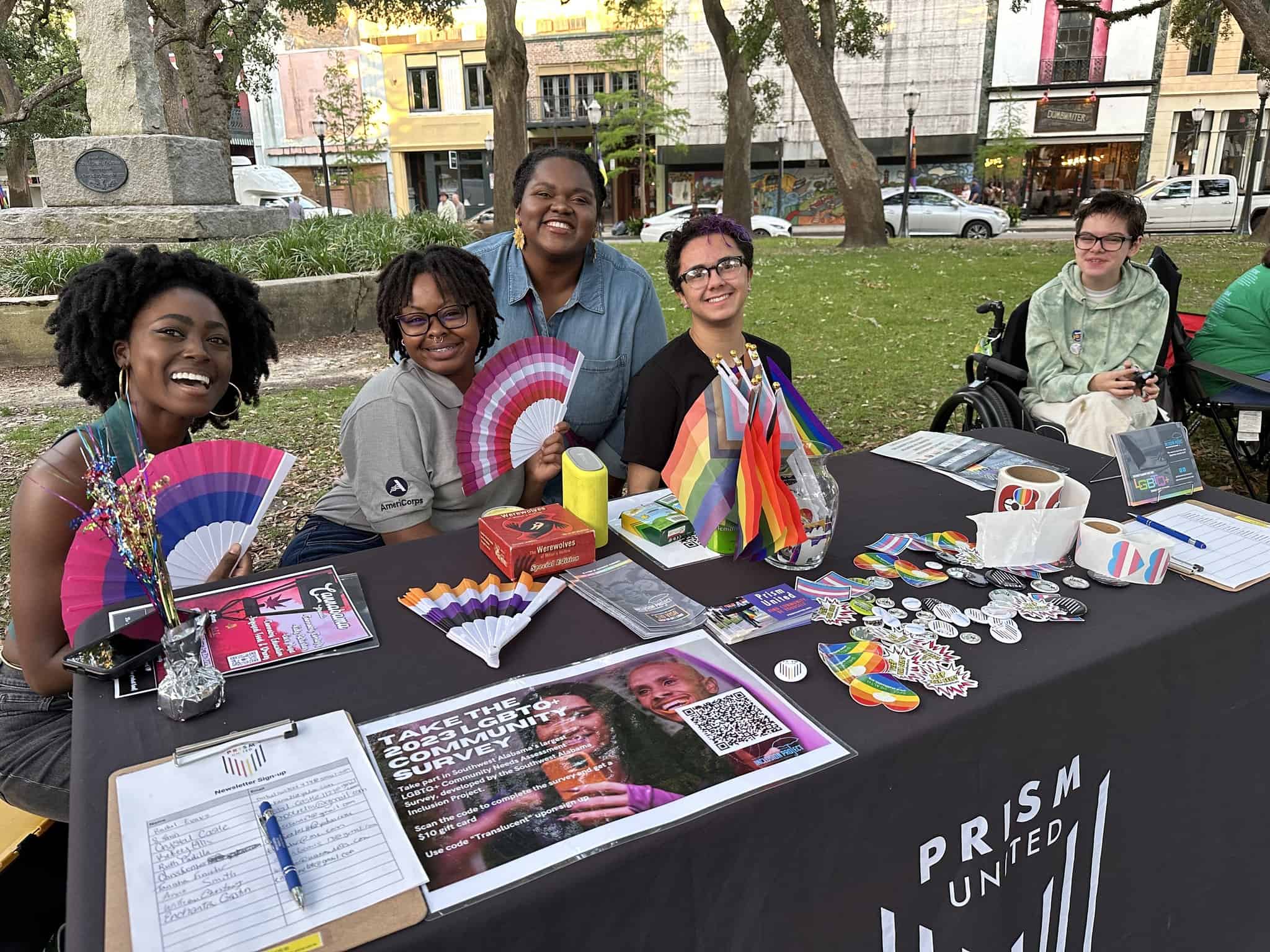 Group of smiling individuals at an outdoor event booth with promotional materials and rainbow flags.