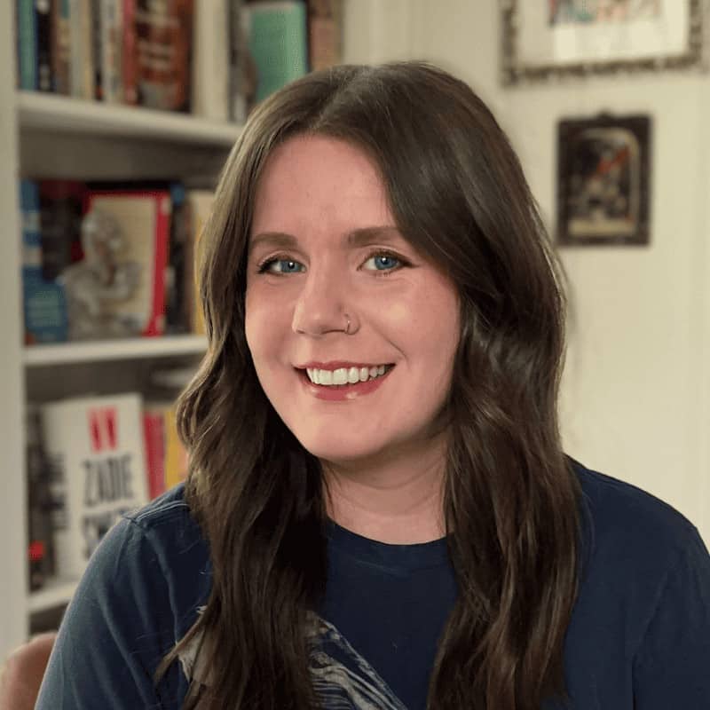 A smiling woman with long brown hair, a nose ring, and a blue top in a room with bookshelves.