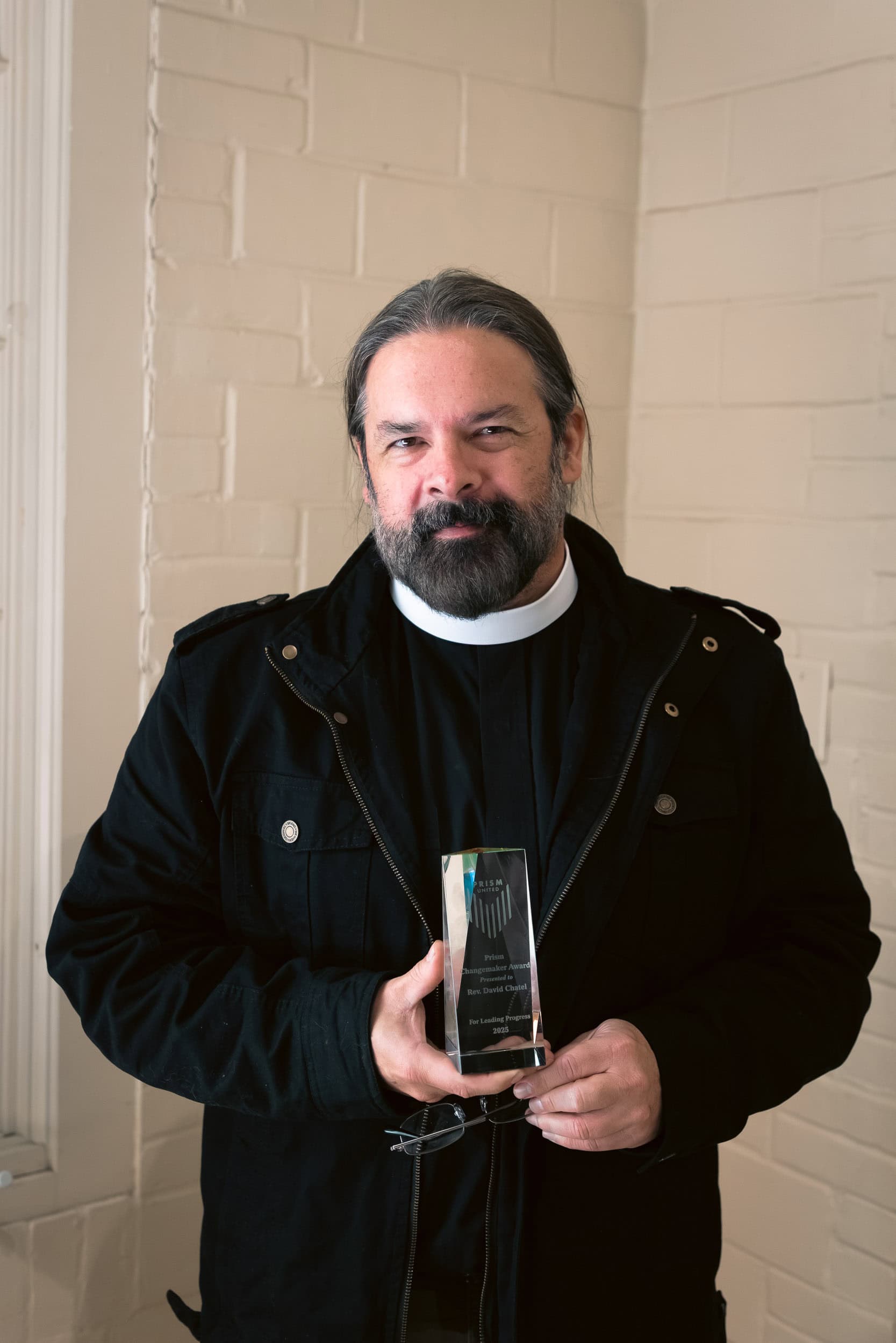RevDaveChatel A man with a beard wearing a black jacket holds a glass award and stands in front of a white brick wall, looking at the camera.