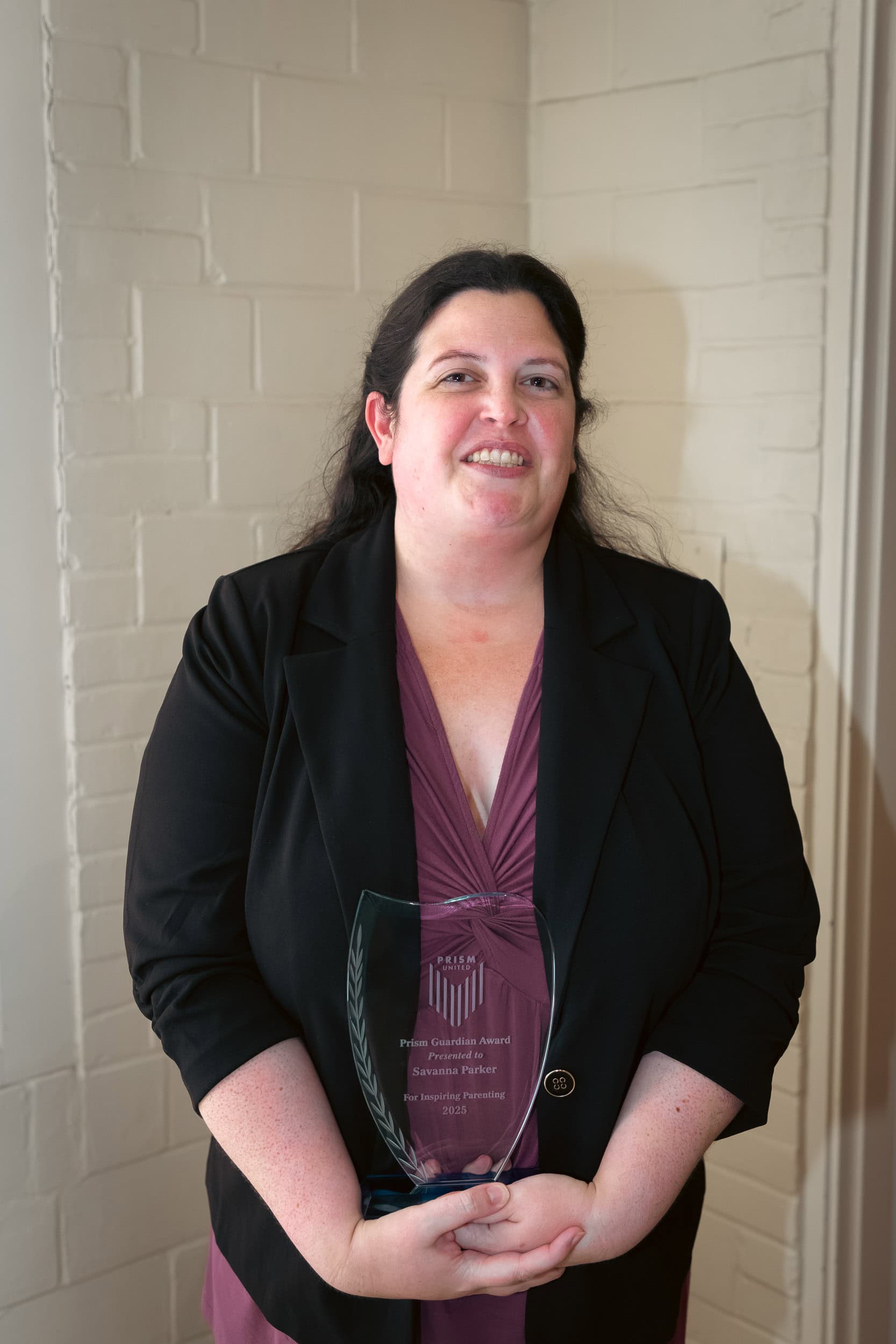 SavannaParker A woman in a maroon dress and black blazer stands indoors, holding a clear glass award and smiling at the camera.