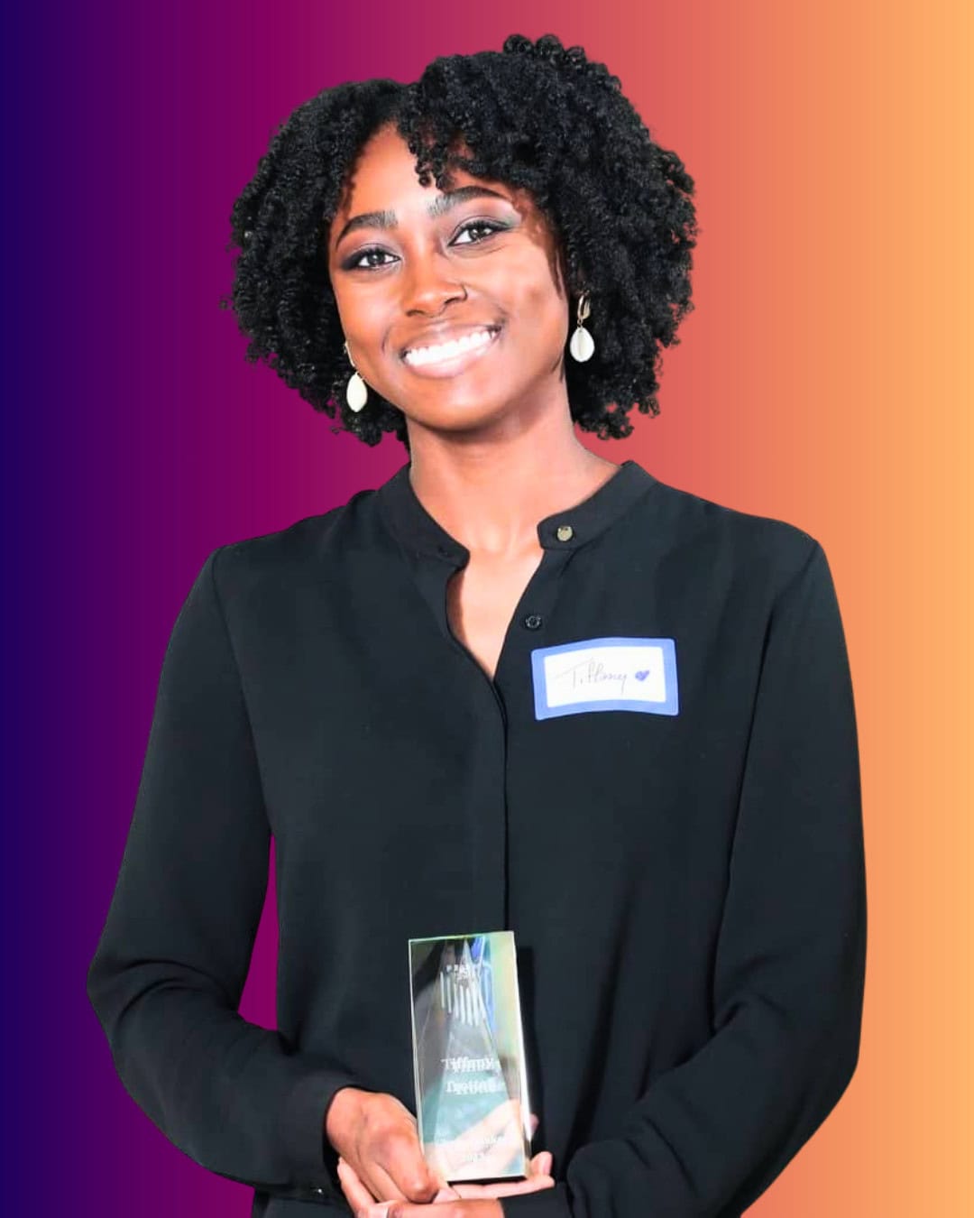 TiffanyTrotter A person with curly hair smiles while holding a glass award. They wear a black shirt and a name tag against a gradient orange and purple background.