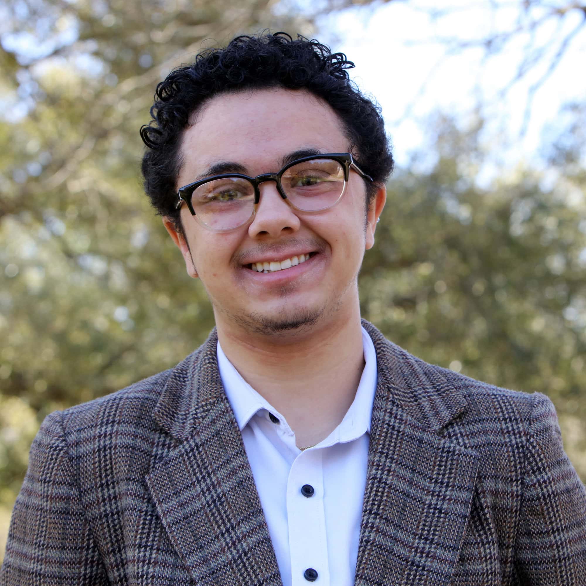 A young man with glasses, curly hair, and a mustache is smiling outdoors while wearing a brown plaid blazer over a white shirt. Trees and sky are in the background.
