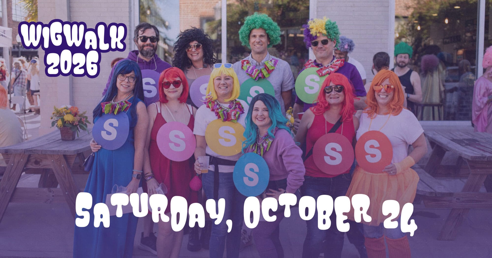 A group of people in colorful wigs and costumes pose outside with signs reading "S," promoting Wig Walk 2026 on Saturday, October 24.