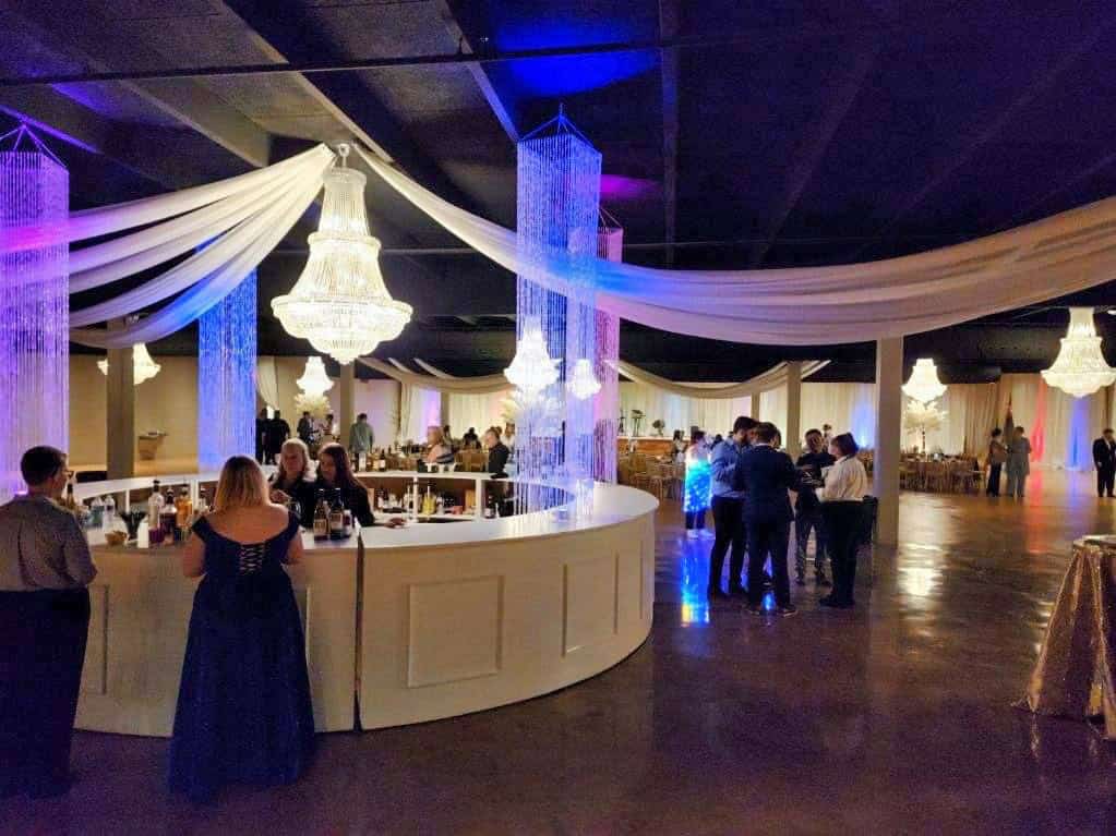 A banquet hall with people gathered around a semi-circular bar. Chandeliers and draped fabric decorate the ceiling. Some patrons are seated and others are standing, mingling near the bar area.