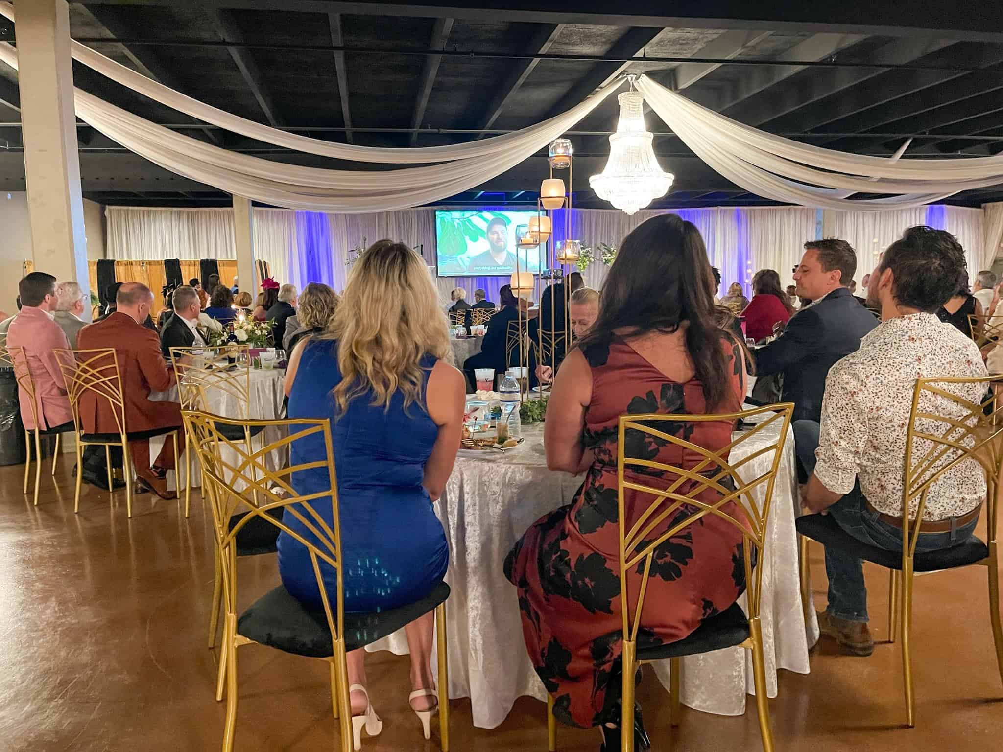 People seated at decorated tables in a banquet hall with draped fabric and a chandelier, watching a presentation on a screen.