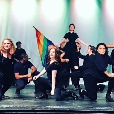 A diverse group of performers on stage, one holding a rainbow flag, with expressions of joy and enthusiasm during a theatrical performance.