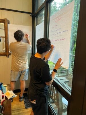 Two individuals write on large sheets of paper taped to a window. One person wears a black shirt and holds the paper, the other wears a gray shirt and writes with a pen. The scene appears to be indoors.