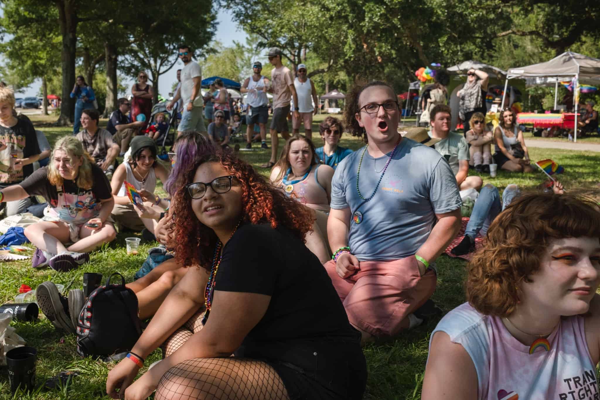 A diverse group of people sitting on grass at an outdoor event, listening attentively, with some holding pride flags.