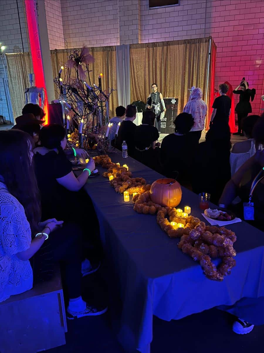 A Halloween-themed event with people sitting at a table decorated with lights, pumpkins, and snacks. A person in costume appears to be performing on stage.