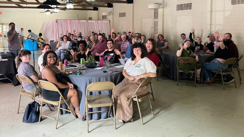 A group of people sitting at tables indoors, smiling and waving at the camera.