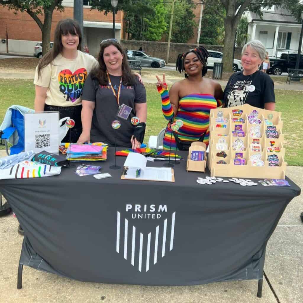 Four people stand behind a table with a "PRISM United" banner, displaying various items, including stickers and buttons, at an outdoor event.