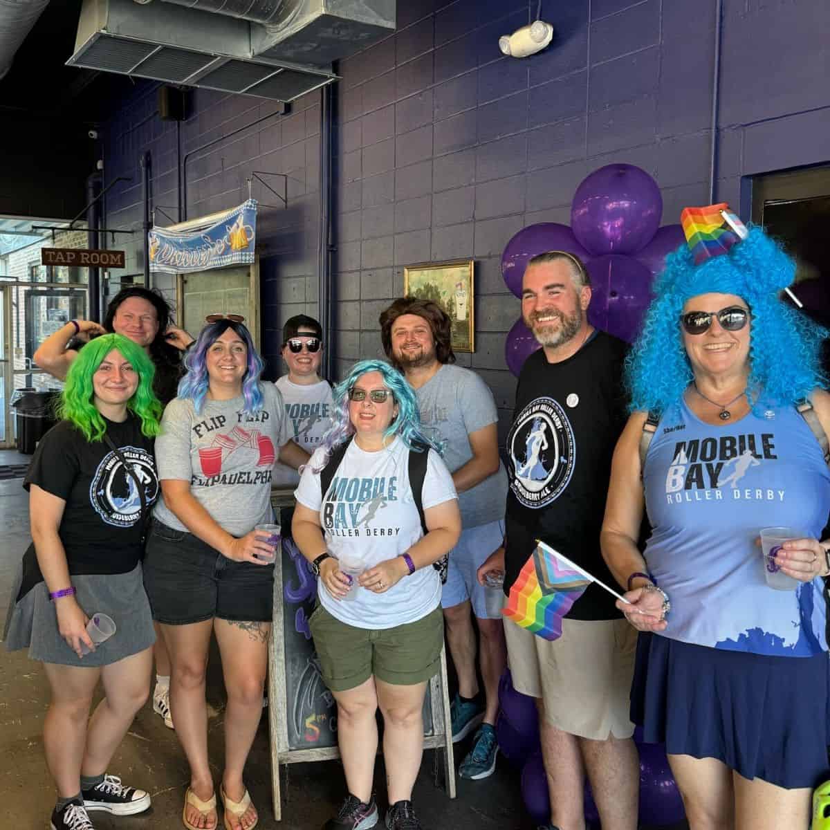 A group of people with colorful hair and outfits stand together indoors, holding small rainbow flags and smiling at the camera.