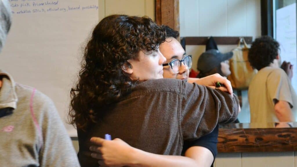 Two people are hugging indoors near a whiteboard. Another person is writing on the whiteboard in the background.