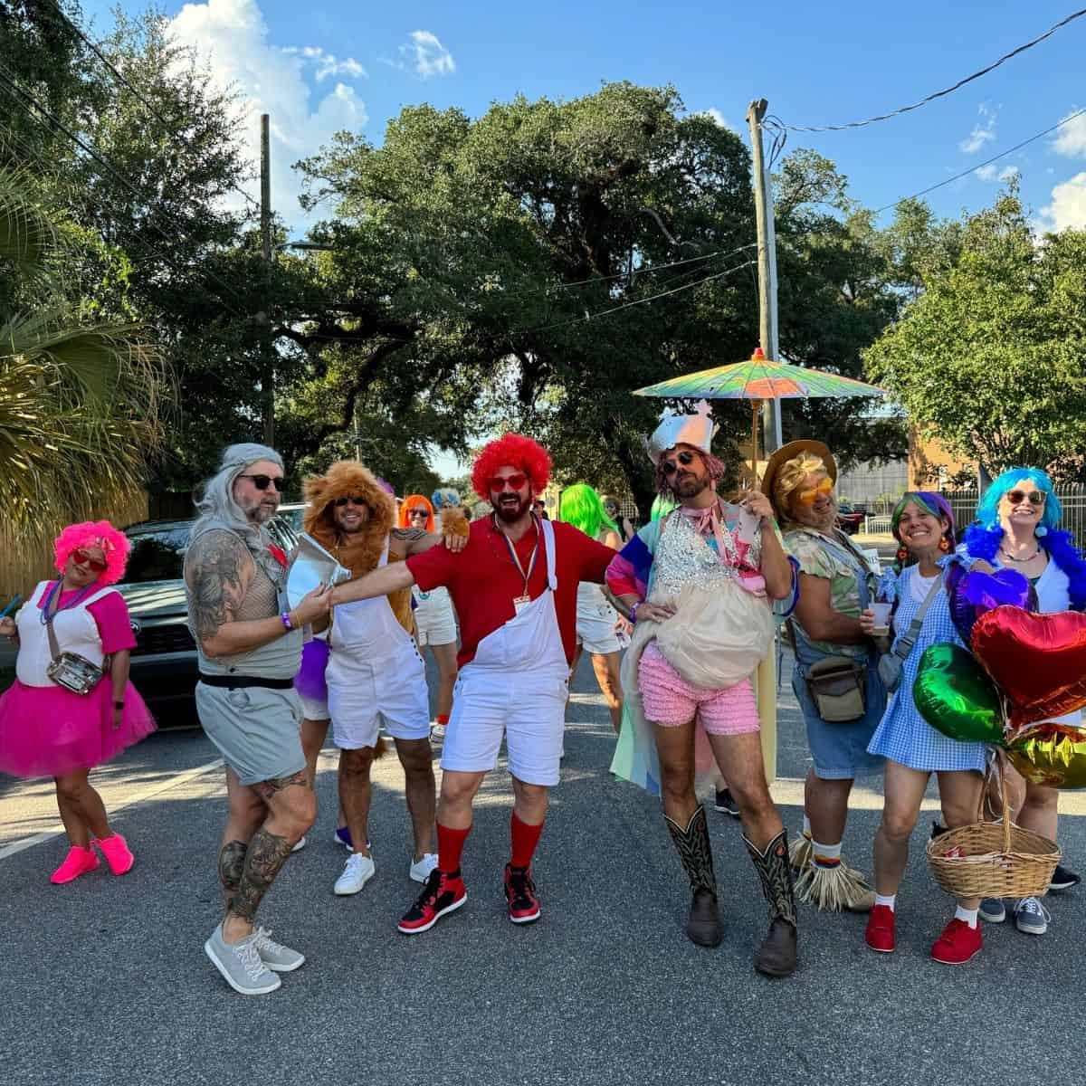 A group of people in colorful, eclectic costumes pose together outdoors, some holding props and wearing wigs. Trees and a clear sky are in the background.