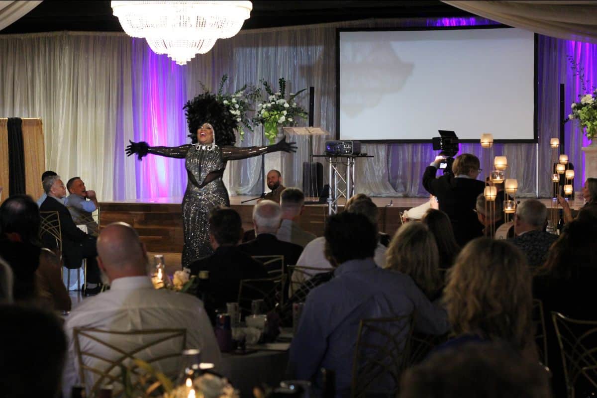 A performer in a glittering outfit and dark gloves with arms outstretched entertains a seated audience in an elegant venue with a large chandelier and a projection screen.