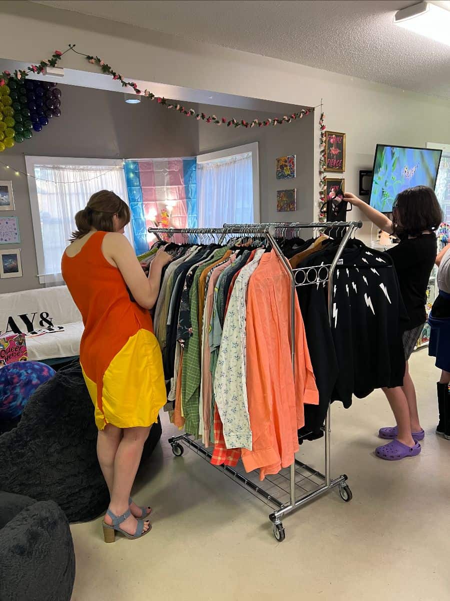 Two people browse a clothing rack filled with colorful shirts and jackets in a decorated room with art on the walls and festive garlands hanging from the ceiling.