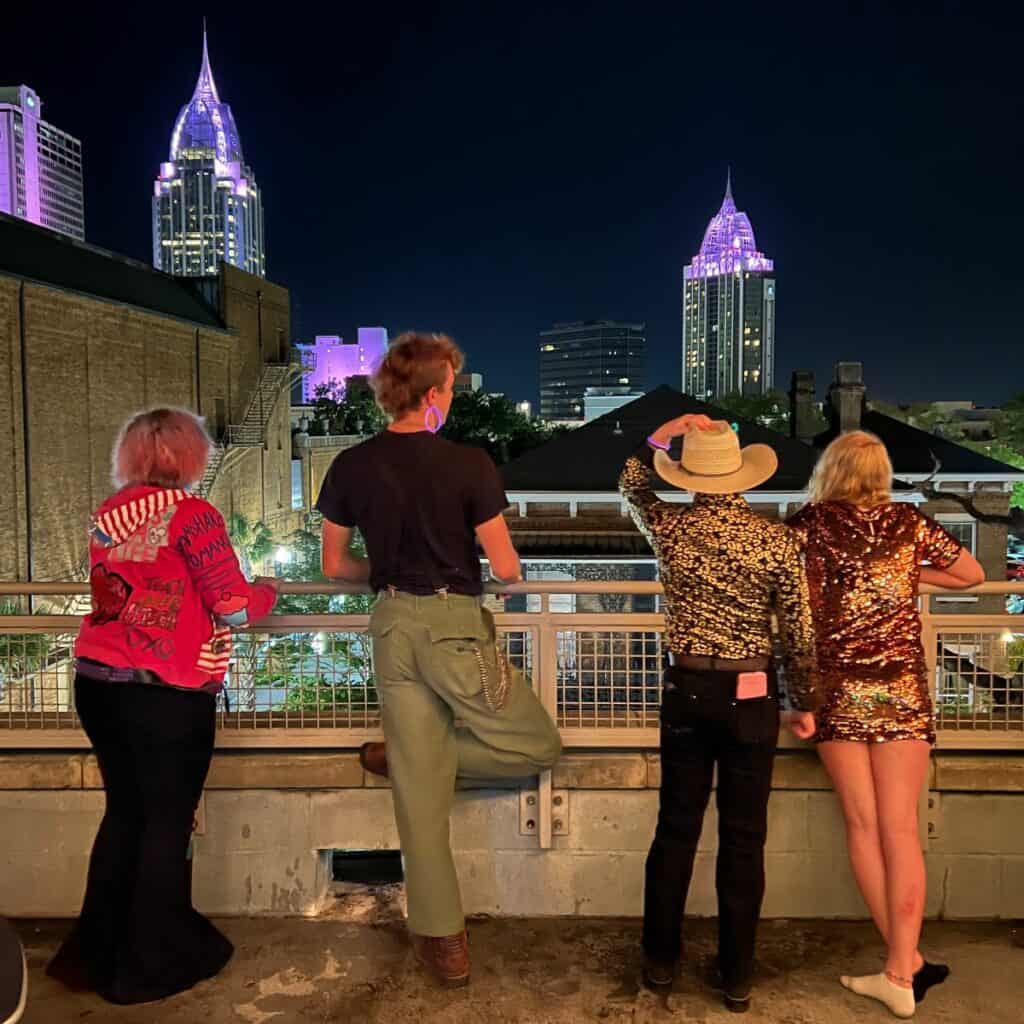 Four people standing on a rooftop at night, observing a city skyline with illuminated buildings.