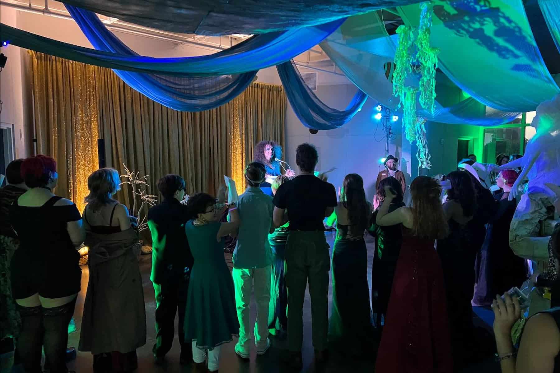 A crowd of people in various costumes and formal wear gather in a dimly lit room with blue and green drapes, watching a performer on stage.