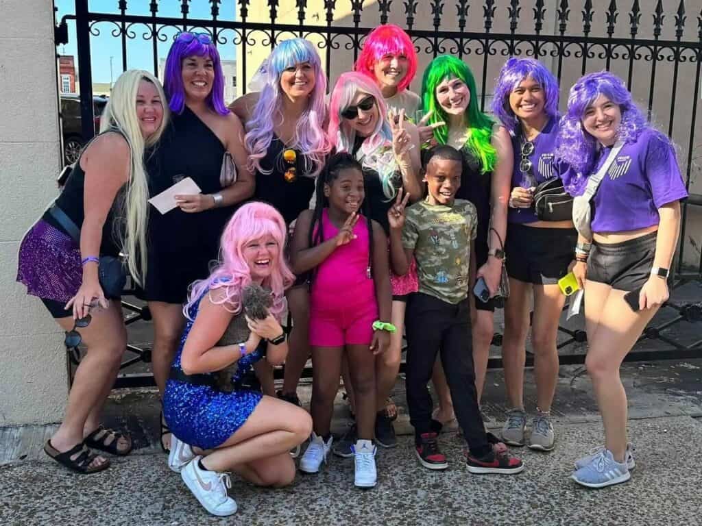 A group of people posing for a photo, many wearing colorful wigs. They are standing in front of a wrought iron fence, smiling and making peace signs. Two young children are included in the group.