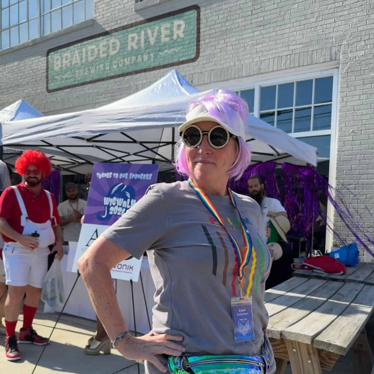 Person with a purple wig and sunglasses stands confidently outside a tent. The event appears to be at Braided River Brewing Company.