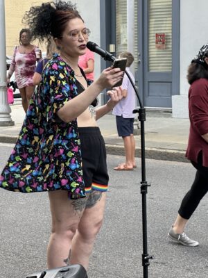 Person with a floral shirt and rainbow shorts reading from a phone into a microphone on a city street. Various pedestrians are visible in the background.