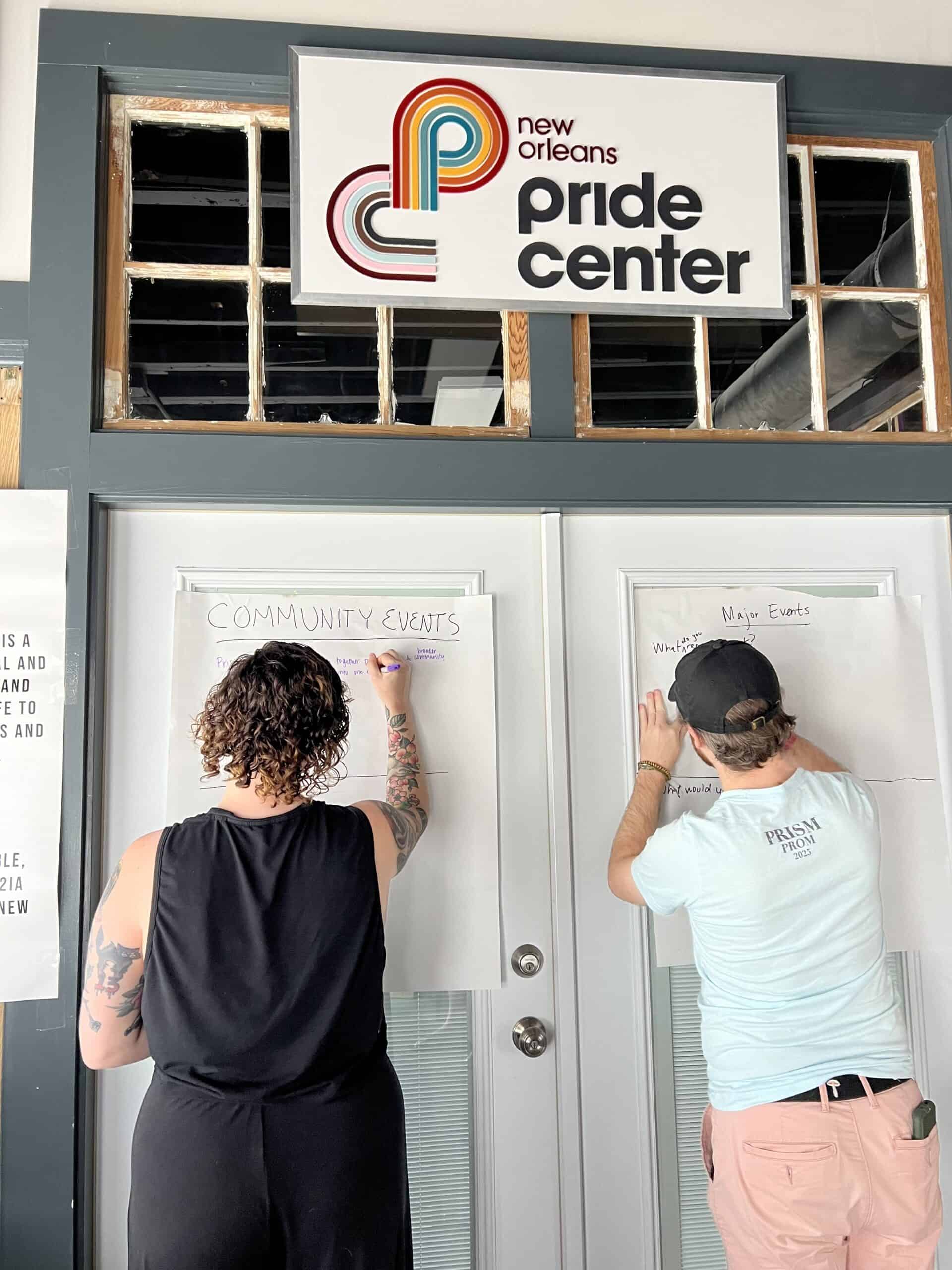 Two people write on large sheets of paper taped to doors under a sign that reads "New Orleans Pride Center.