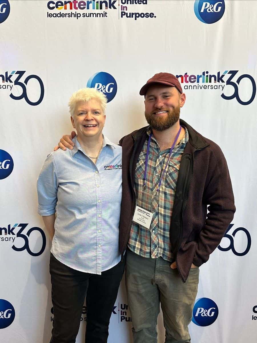 Two people stand smiling in front of a backdrop with Centerlink and P&G logos. The person on the left has short white hair and is wearing a light blue shirt; the person on the right has a beard and is wearing a cap.