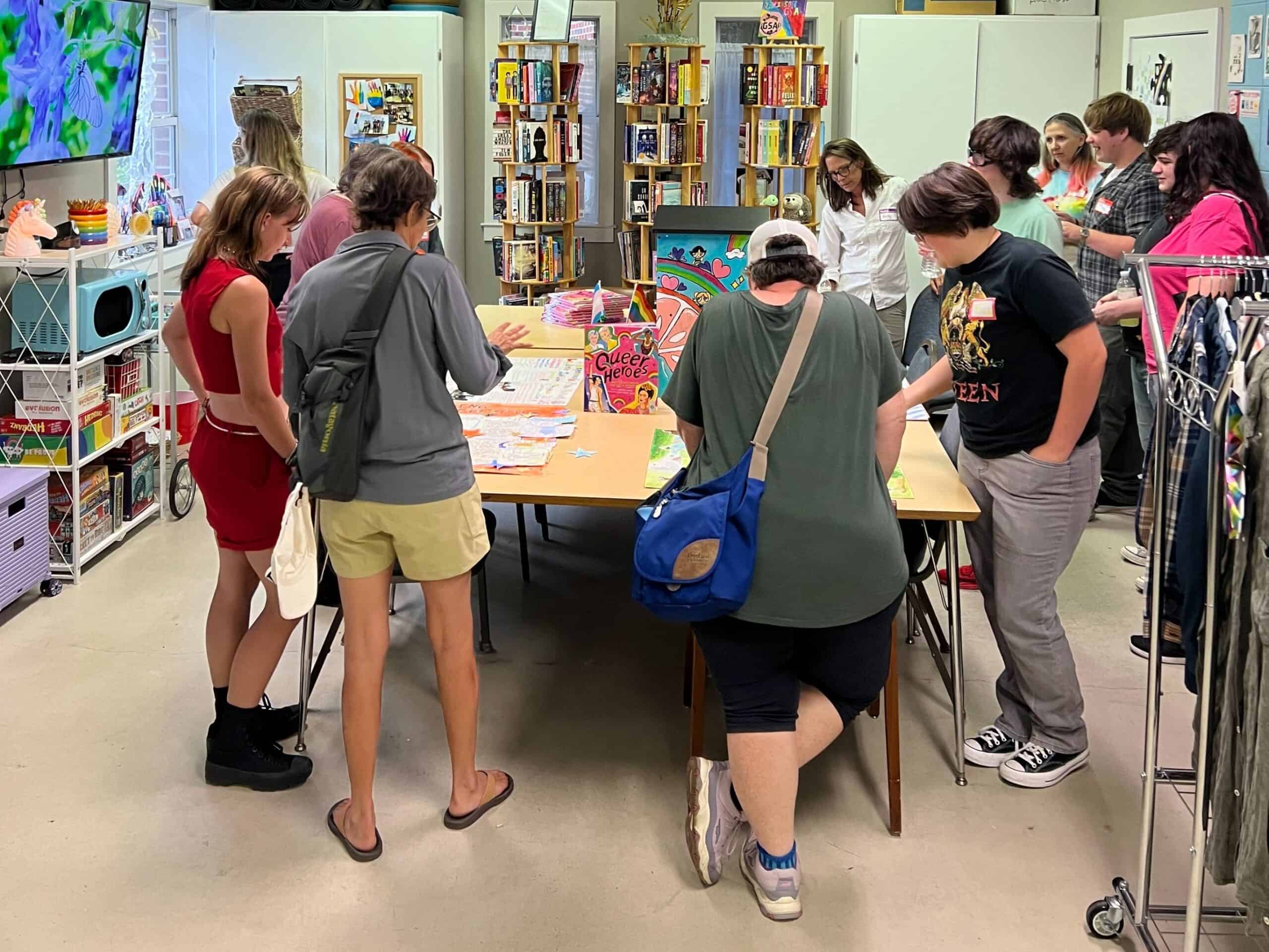 A group of people stand around a table engaged in activities, with bookshelves and various items visible in the background of a brightly lit room.