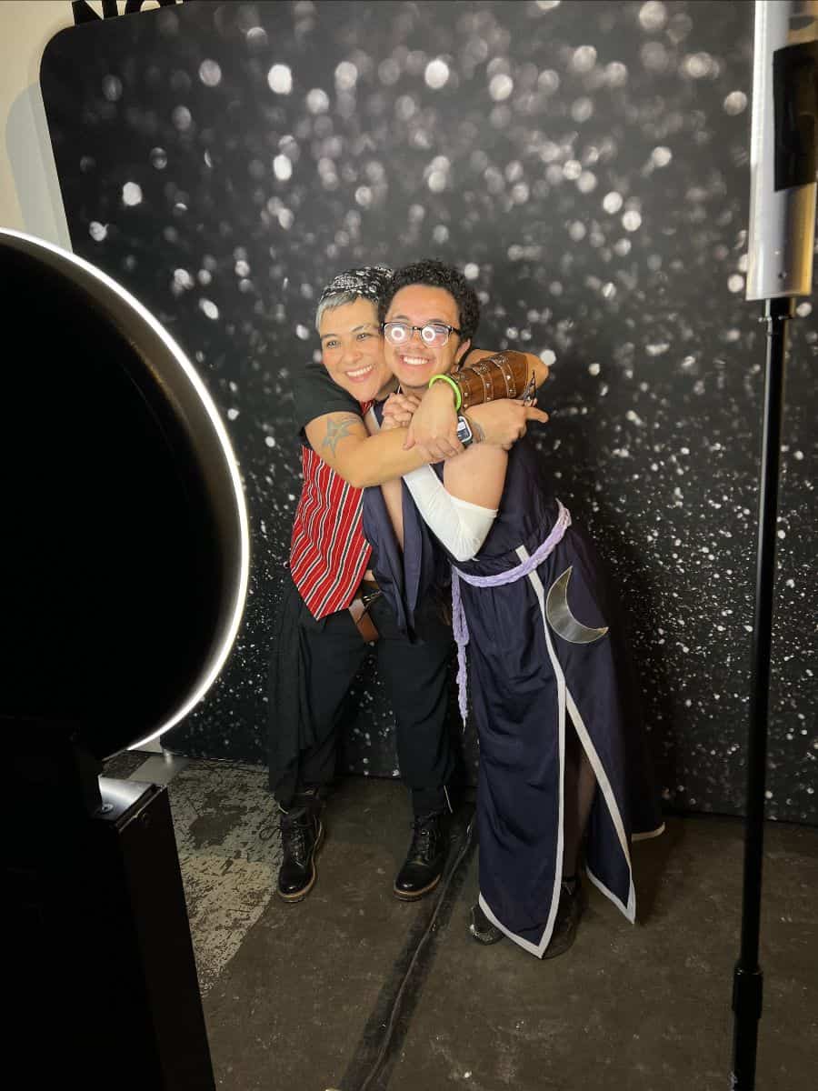 Two people embracing and smiling in front of a sparkly backdrop, one wearing a striped shirt and the other in a costume with a crescent moon design.