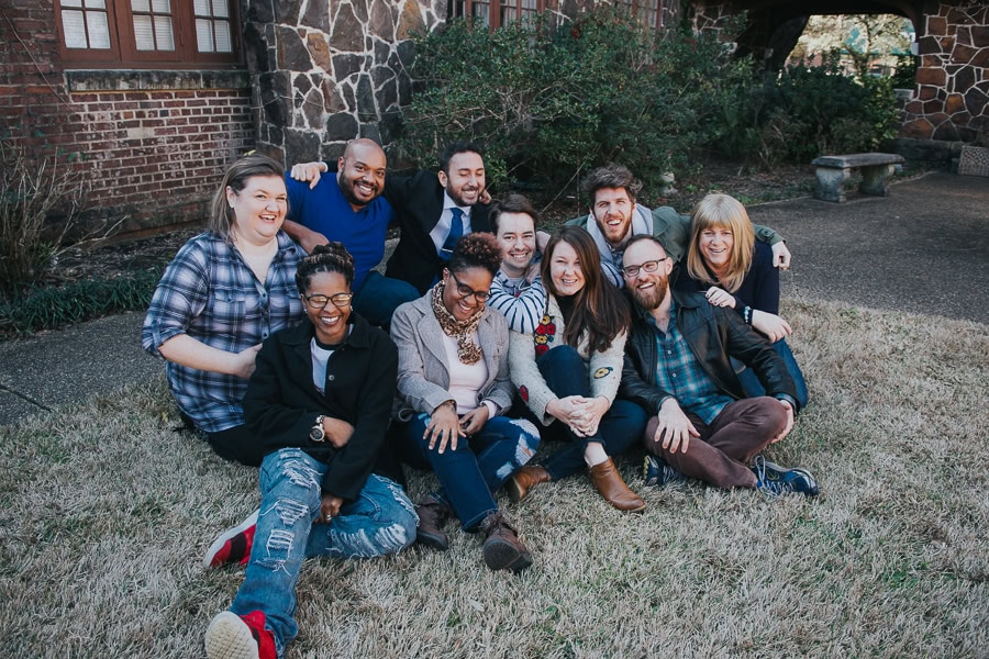 A group of eleven adults pose together, smiling, while sitting and kneeling on grass in front of a stone building.