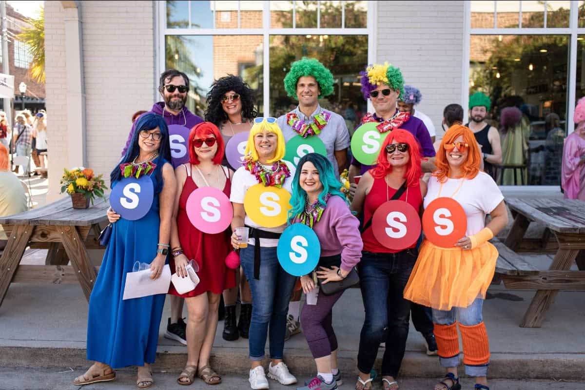 A group of eleven people in colorful wigs and costumes stand outside holding large paper S’s, posing for a group photo in front of a building with large windows.